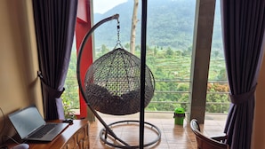 Una habitación con una laptop sobre un escritorio, una silla colgante, una mesita con una botella verde y vistas a campos escalonados a través de un ventanal grande.