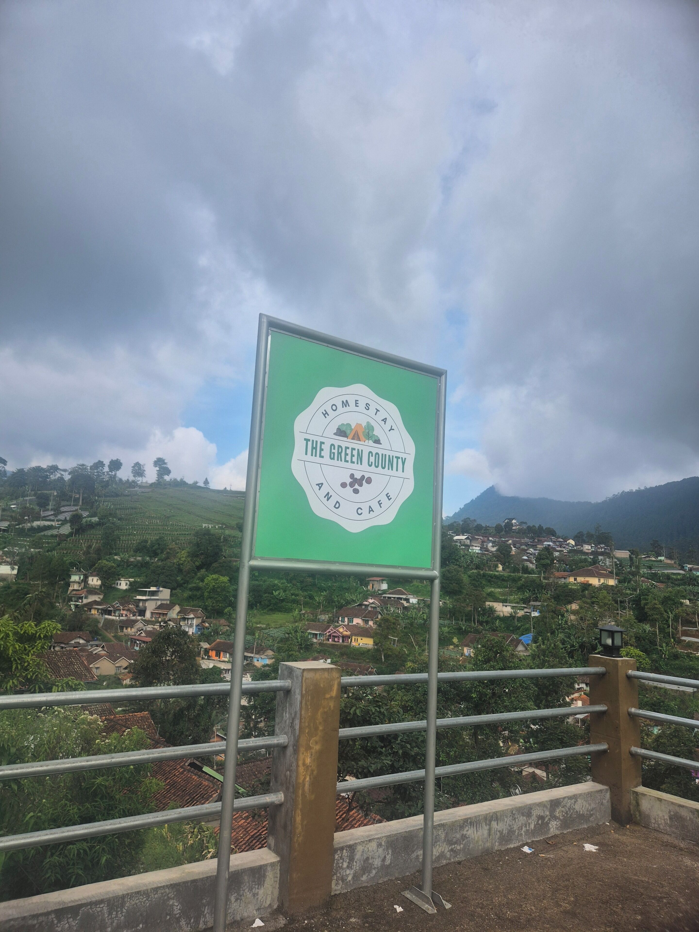 Un letrero con el texto 'Homestay The Green County and Cafe', rodeado de un paisaje rural con casas y colinas.