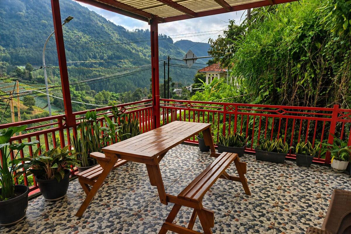 A covered outdoor area with a wooden table and benches, surrounded by potted plants and a red railing.