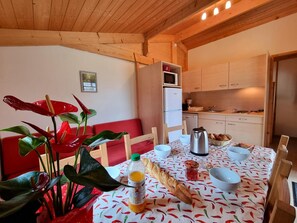 A cozy kitchen with wooden cabinets, a microwave, and a refrigerator. There is a dining table with a patterned tablecloth, chairs, and a red bench. A plant with red leaves is on the table, and a bottle of orange juice is visible. - Chalet in nature, pets welcome - 6 guests (Usson-en-Forez)