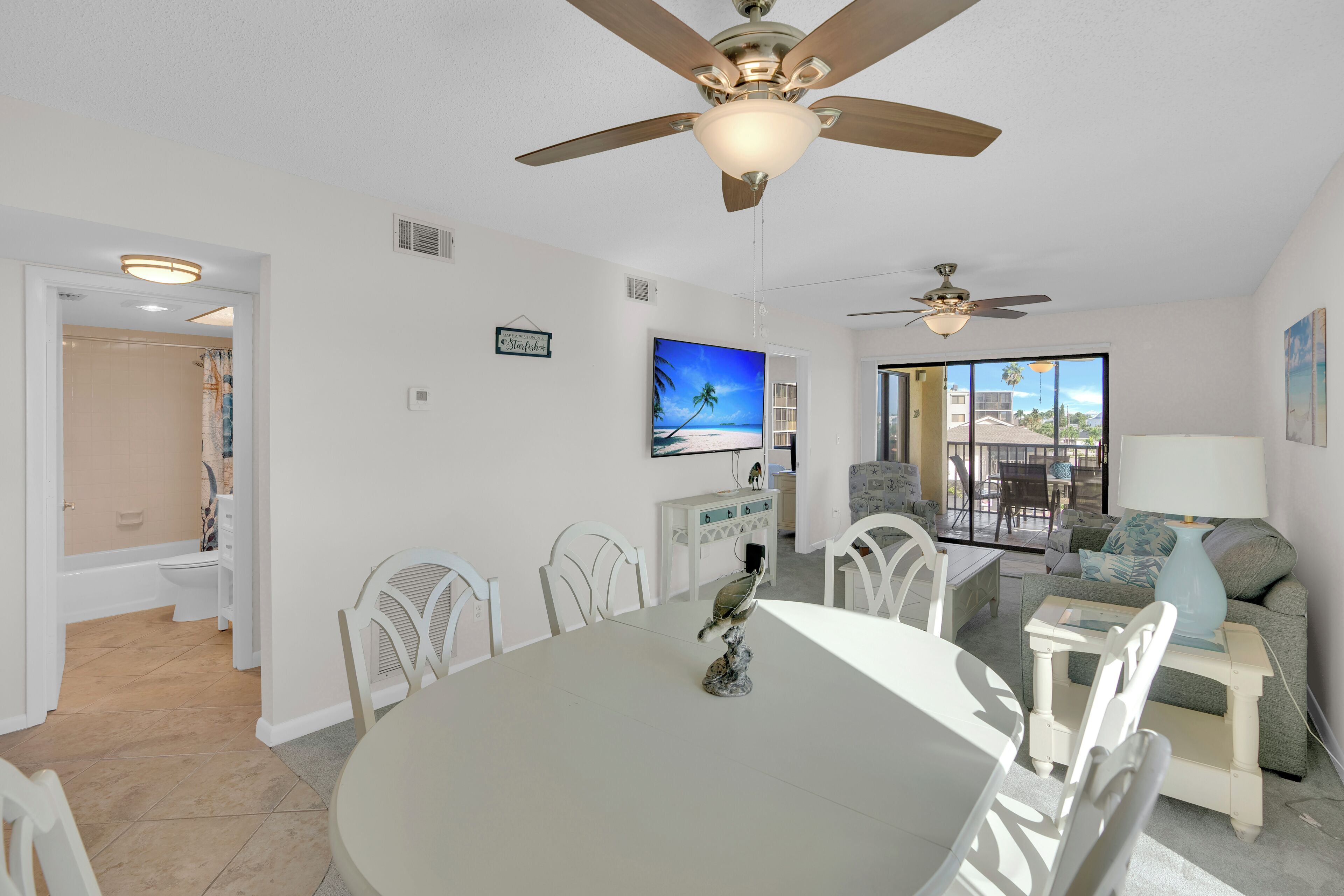 A modern living room with a dining area, a ceiling fan, a flat-screen TV, and a view through large windows.