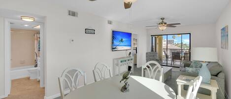 A modern living room with a dining area, a ceiling fan, a flat-screen TV, and a view through large windows.