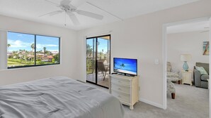 Een slaapkamer met een bed, een televisie, een dressoir en een balkon met uitzicht op palmbomen en huizen.