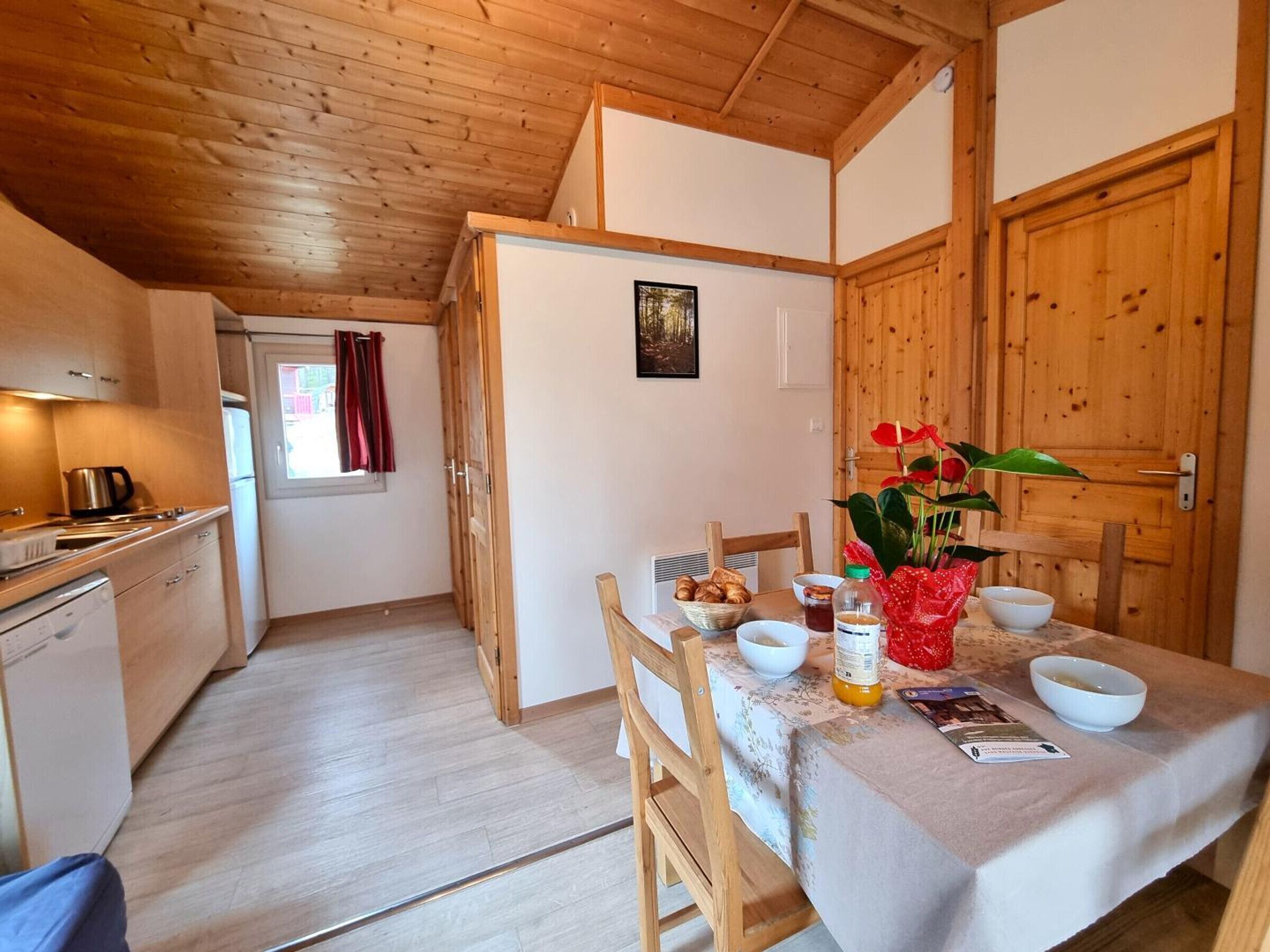 A compact kitchen with a dining area, wooden ceiling, and a small window with curtains.