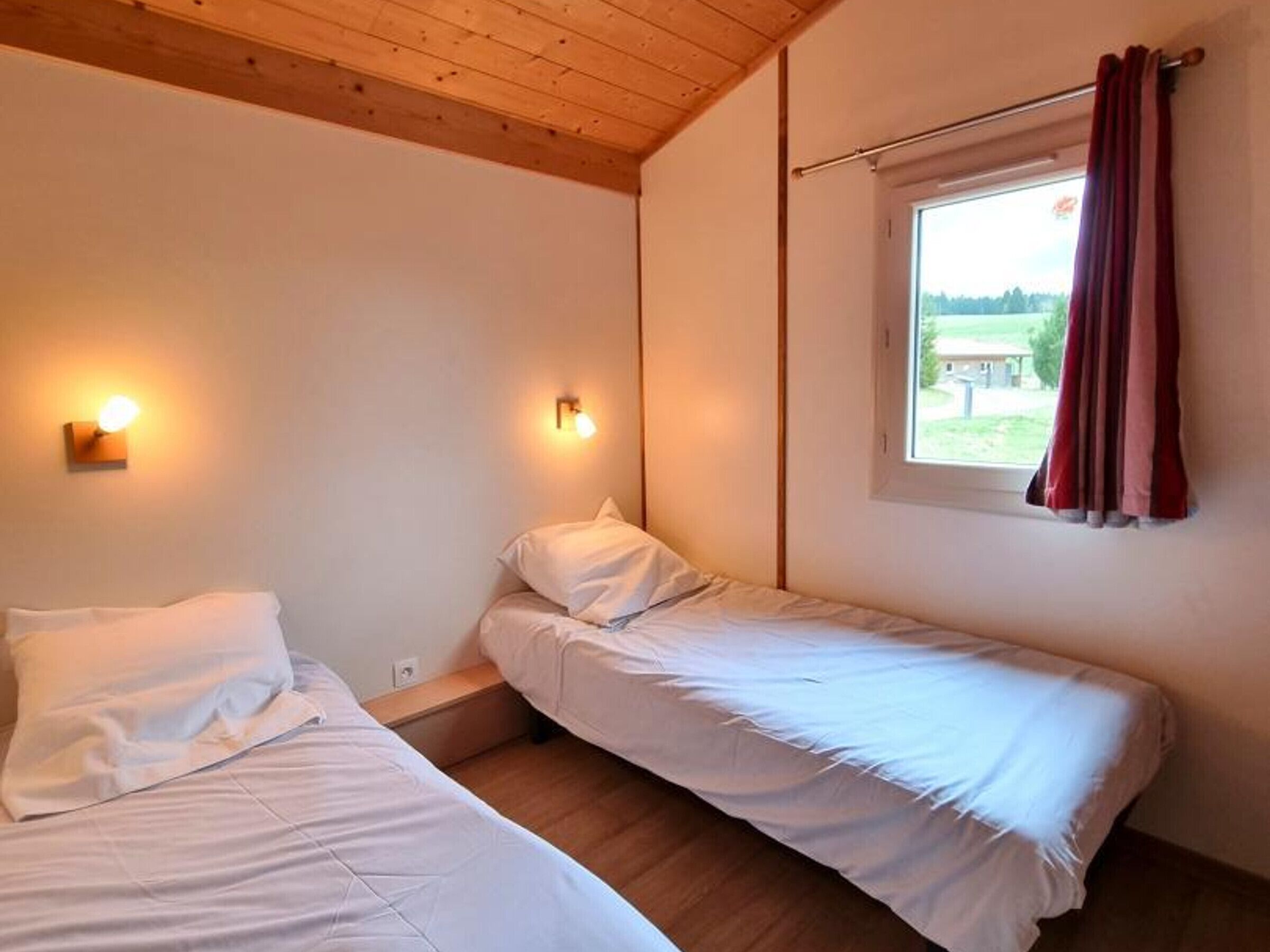A room with two single beds, wooden ceiling, two wall-mounted lights, a window with curtains, and a view of greenery outside.