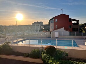 A swimming pool with a fence, surrounded by greenery and a building with a red facade. - Apartment marina view, pool - 4 guests (Saint-Cyprien)