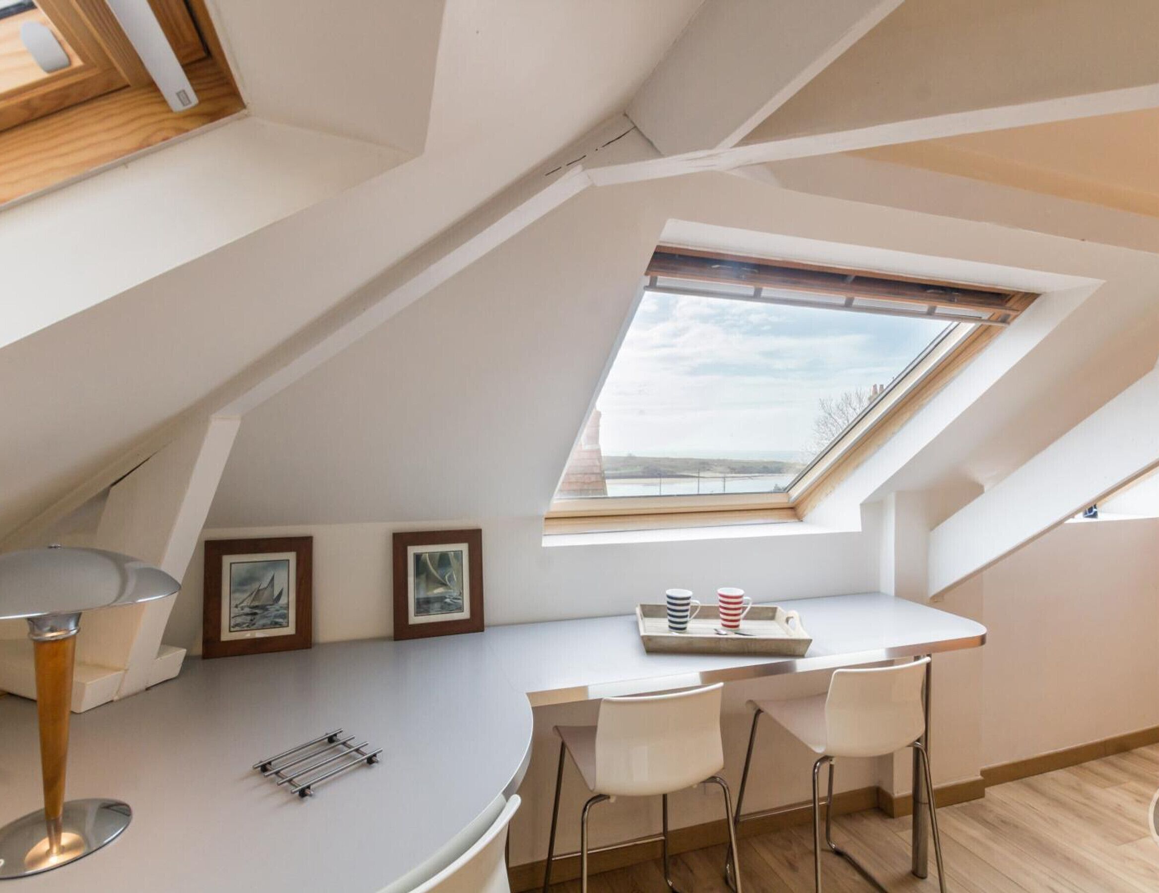 Une chambre de style loft moderne, dotée d’une lucarne, d’un bureau blanc aux lignes courbes, de deux chaises blanches et de tableaux encadrés accrochés au mur.