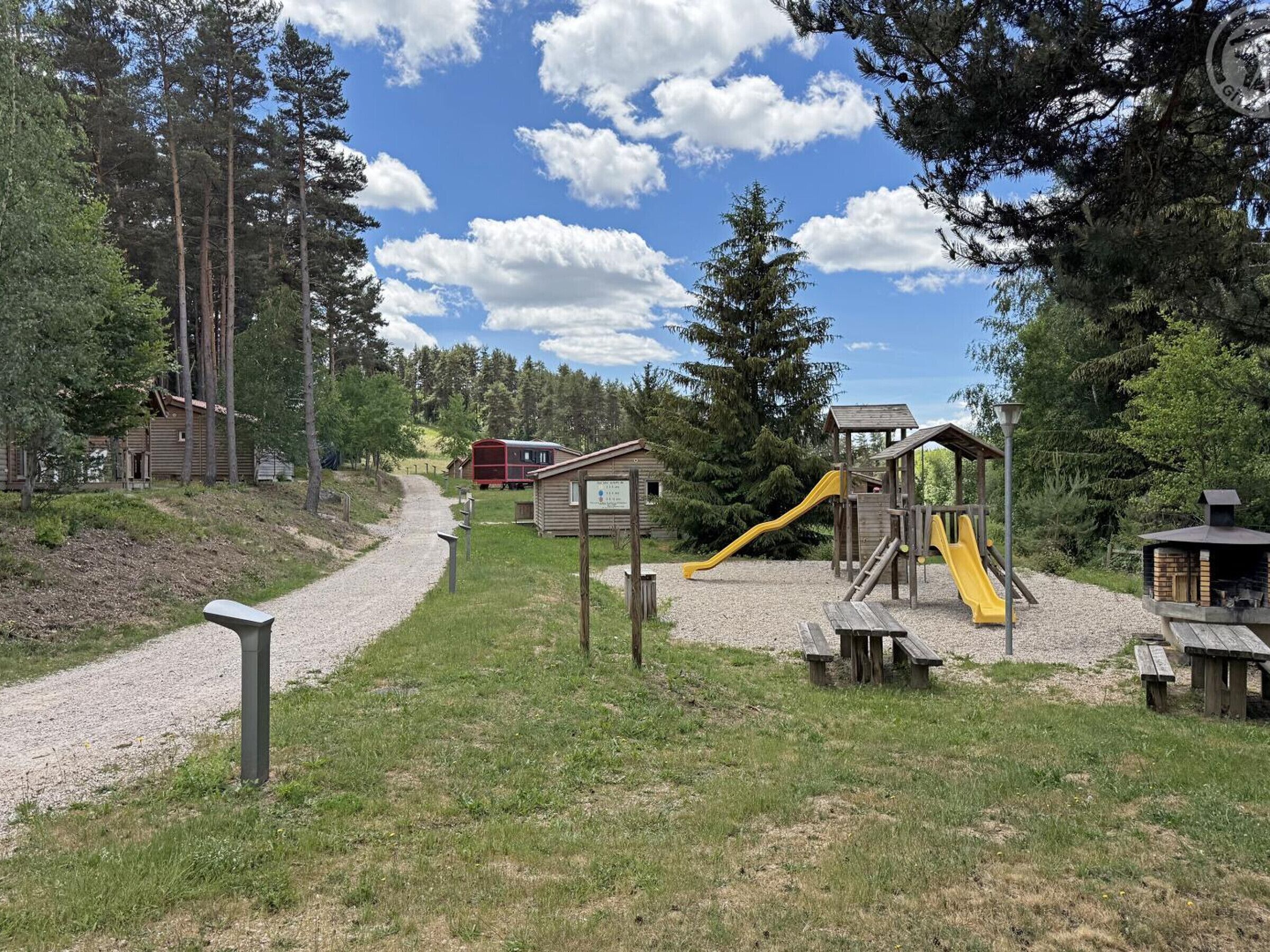Un parque infantil con tobogán y estructuras para escalar, rodeado de árboles y un camino de grava.