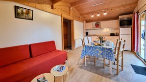 Un ambiente accogliente con un divano rosso, un tavolo da pranzo con sedie, un tavolino con riviste, travi in legno a soffitto, un angolo cottura con pensili ed elettrodomestici, e una finestra con tende.