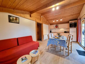A cozy room with a red sofa, a dining table with chairs, a small table with magazines, wooden ceiling beams, a kitchen area with cabinets and appliances, and a window with curtains.