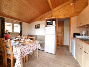 Eine kleine Küche mit Holdekor, einer Mikrowelle und einem Kühlschrank. Ein Esstisch mit Stühlen und ein kleines Fenster mit Vorhängen sind ebenfalls vorhanden.