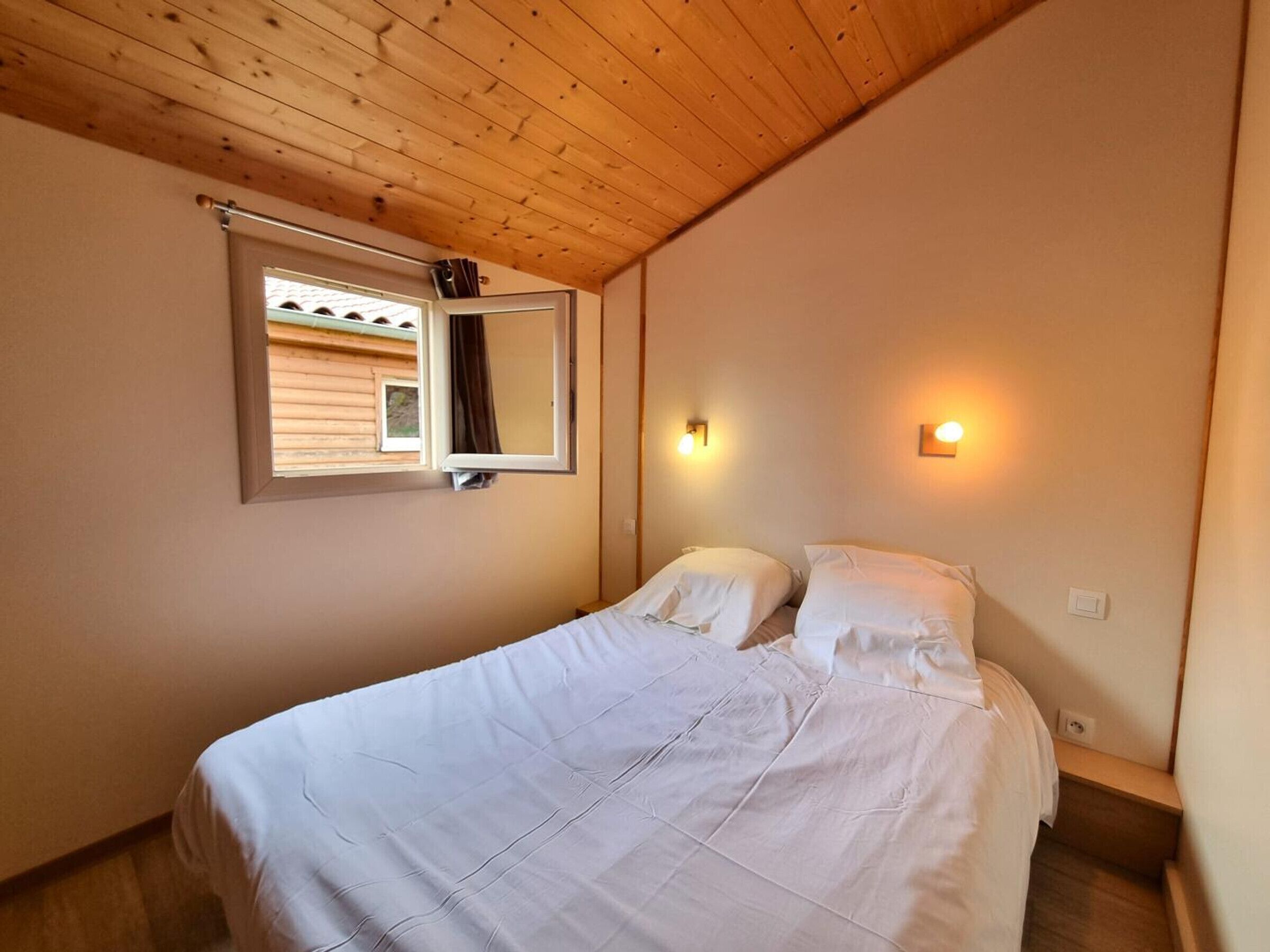 Un dormitorio con techo de madera, una cama con ropa de cama blanca, una ventana con vista a un edificio y luces fijadas en la pared.