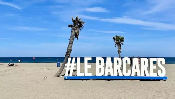 Una platja amb palmeres i un cartell amb la inscripció '#LE BARCARES'.