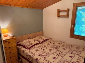 A bedroom with a wooden headboard, a bed with patterned bedding, a nightstand with a lamp, a dresser, and a window with a view of trees. - Cozy studio with mountain view - 4 guests (Demi-Quartier)