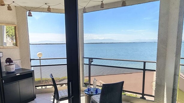 En balkong med utsikt över en sjö, ett bord med stolar och ett litet skåp.