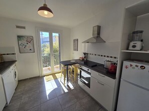 A kitchen with a dining table, chairs, a stove, a refrigerator, and a hanging light fixture. - Bright apartment near town center - 5 guests (Calvi)