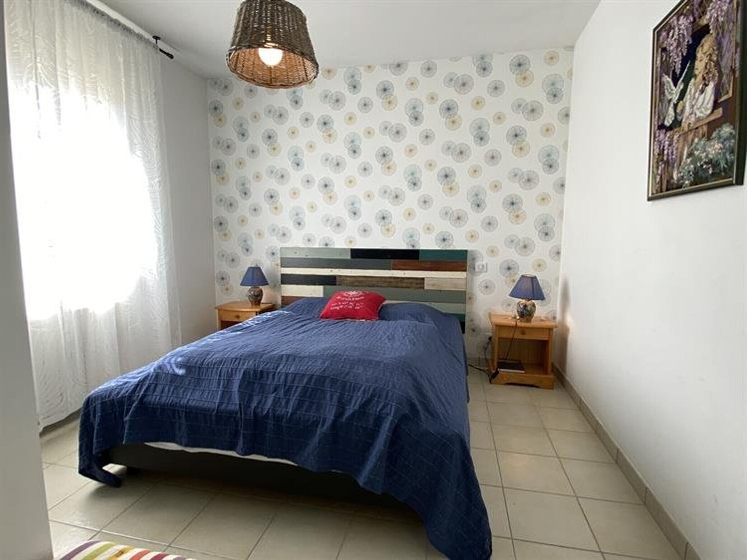 A bedroom with a bed, two bedside tables, a hanging light fixture, a window with curtains, and a wall with a floral pattern.