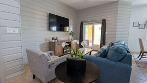Ein Wohnzimmer mit grauem Sofa, grauem Sessel, Couchtisch, flachbildschirmfernseher an der Wand und einer Glastür zum Balkon.