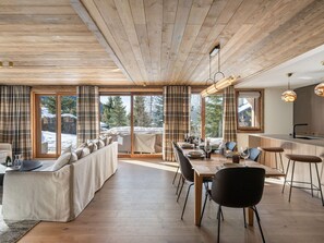 Ein modernes Interieur mit Holzbalkendecke, einem Essbereich mit langem Tisch und Stühlen, einer Couch und großen Fenstern mit Vorhängen.