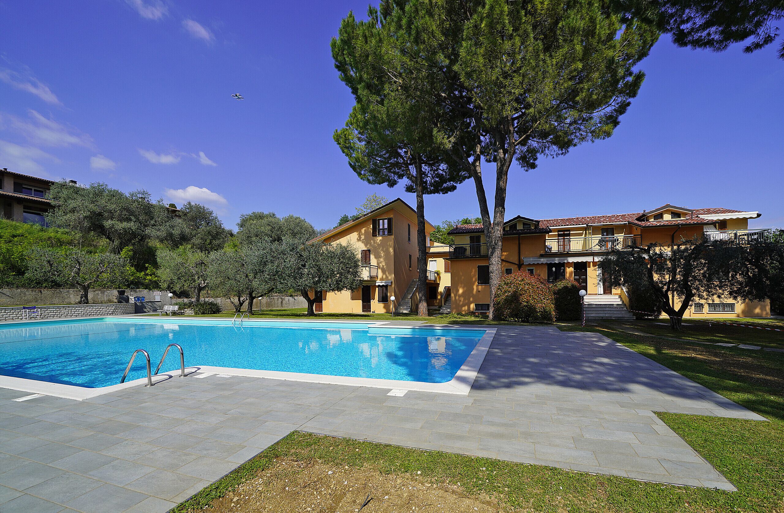 A swimming pool with a clear blue sky, a large tree, and a yellow building with balconies.