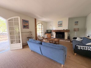 Ein Wohnzimmer mit Sofa, Sesseln, Kamin, Fernseher und einer zur Außenseite führenden Tür.