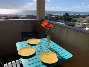 A balcony with a table set for two, a vase with flowers, and a view of the sea and buildings. - Apartment sea view, parking - 6 guests (Fleury-Saint-Pierre-la-Mer)