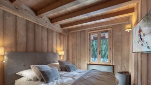Ein Schlafzimmer in einer Holzhütte mit einem Bett, Kissen, einem Nachttisch, einer Lampe und einem Fenster mit Blick auf Bäume.