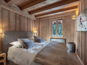 Ein Schlafzimmer in einer Holzhütte mit einem Bett, Kissen, einem Nachttisch, einer Lampe und einem Fenster mit Blick auf Bäume.