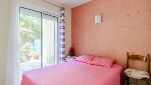 A bedroom with a large bed, a nightstand, a chair, a window with curtains, and a view of greenery outside. - Ground floor apartment near beach - 4 guests (Fleury-Saint-Pierre-la-Mer)