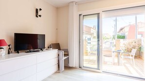 A modern living room with a flat-screen TV, a white cabinet, a red lamp, and a sliding glass door leading to an outdoor patio area. - Ground floor apartment near beach - 4 guests (Fleury-Saint-Pierre-la-Mer)