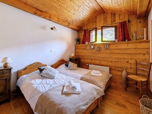 A wooden cabin bedroom with two beds, a window with red curtains, a radiator, and a wooden ceiling.