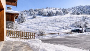 A snow-covered landscape with a wooden building, stone pathway, and a road with parked cars.