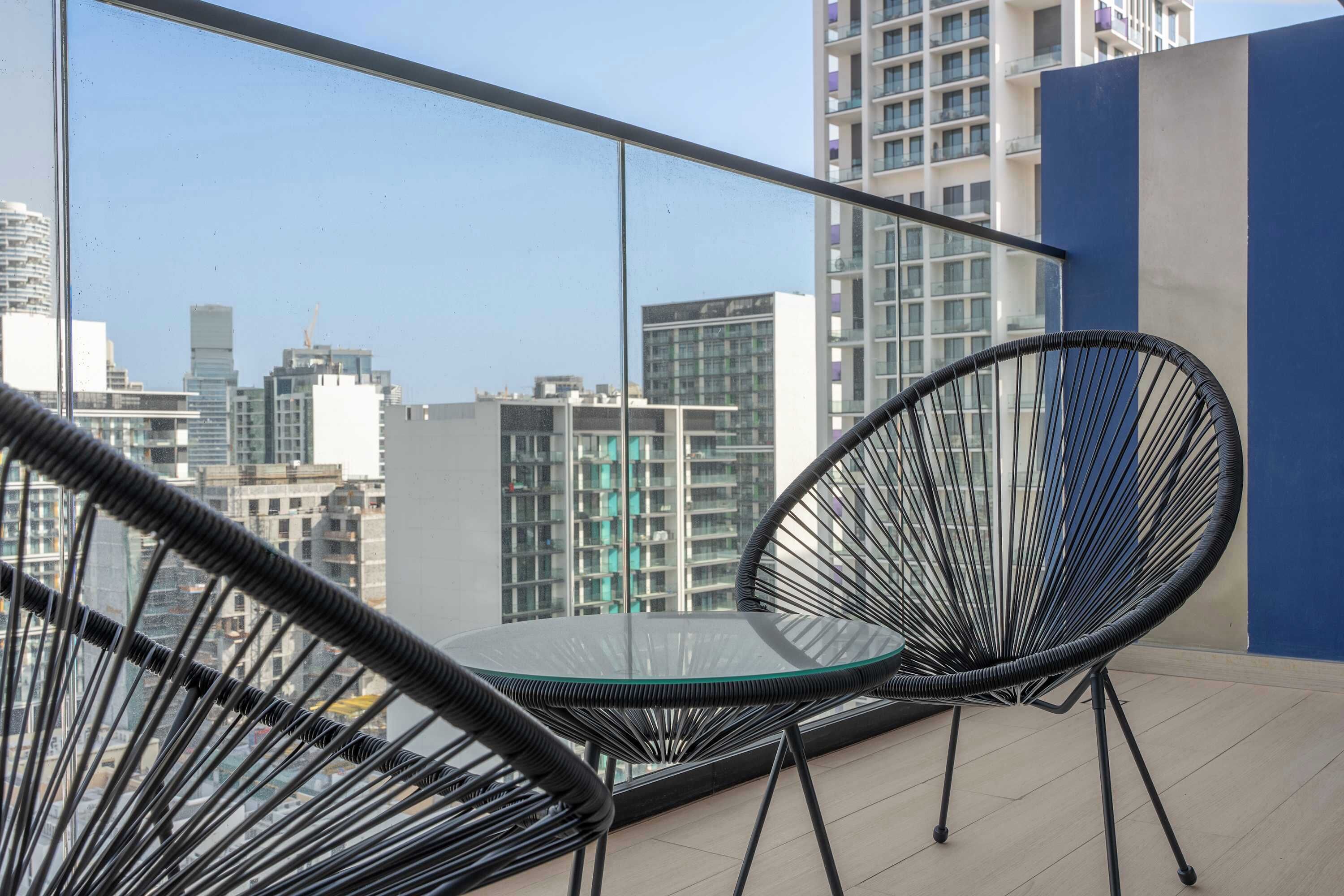 Un balcon avec une table en verre et deux chaises en fil de fer noir, donnant sur un paysage urbain avec des bâtiments modernes.
