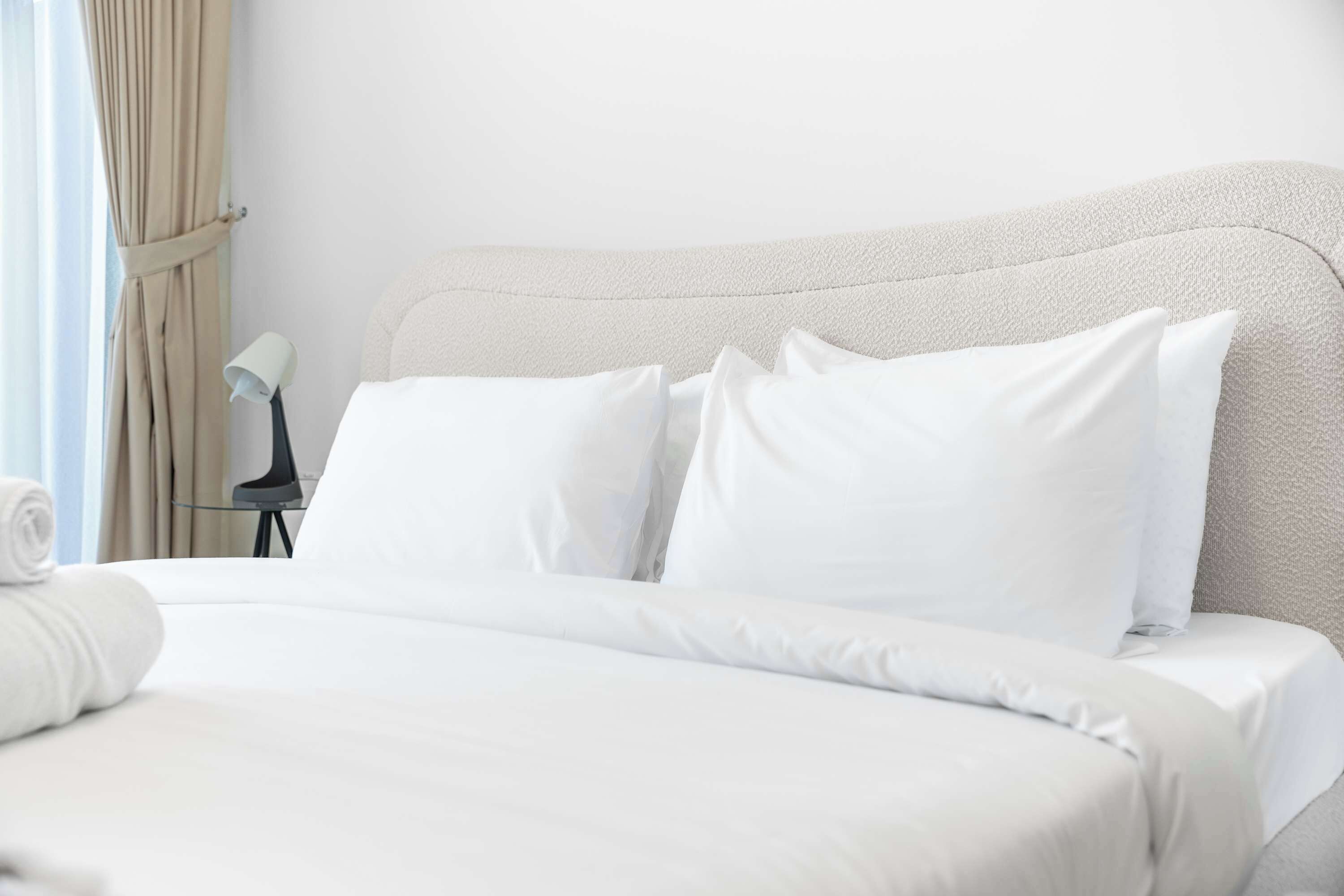 Een net opgemaakt bed met witte lakens en kussens, een klein nachtkastje met een lamp, en een gedeeltelijk getrokken gordijn.