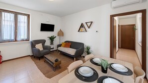 Un saló amb un sofà, una taula de dinar, un televisor fixat a la paret i una porta que dona a una altra habitació.