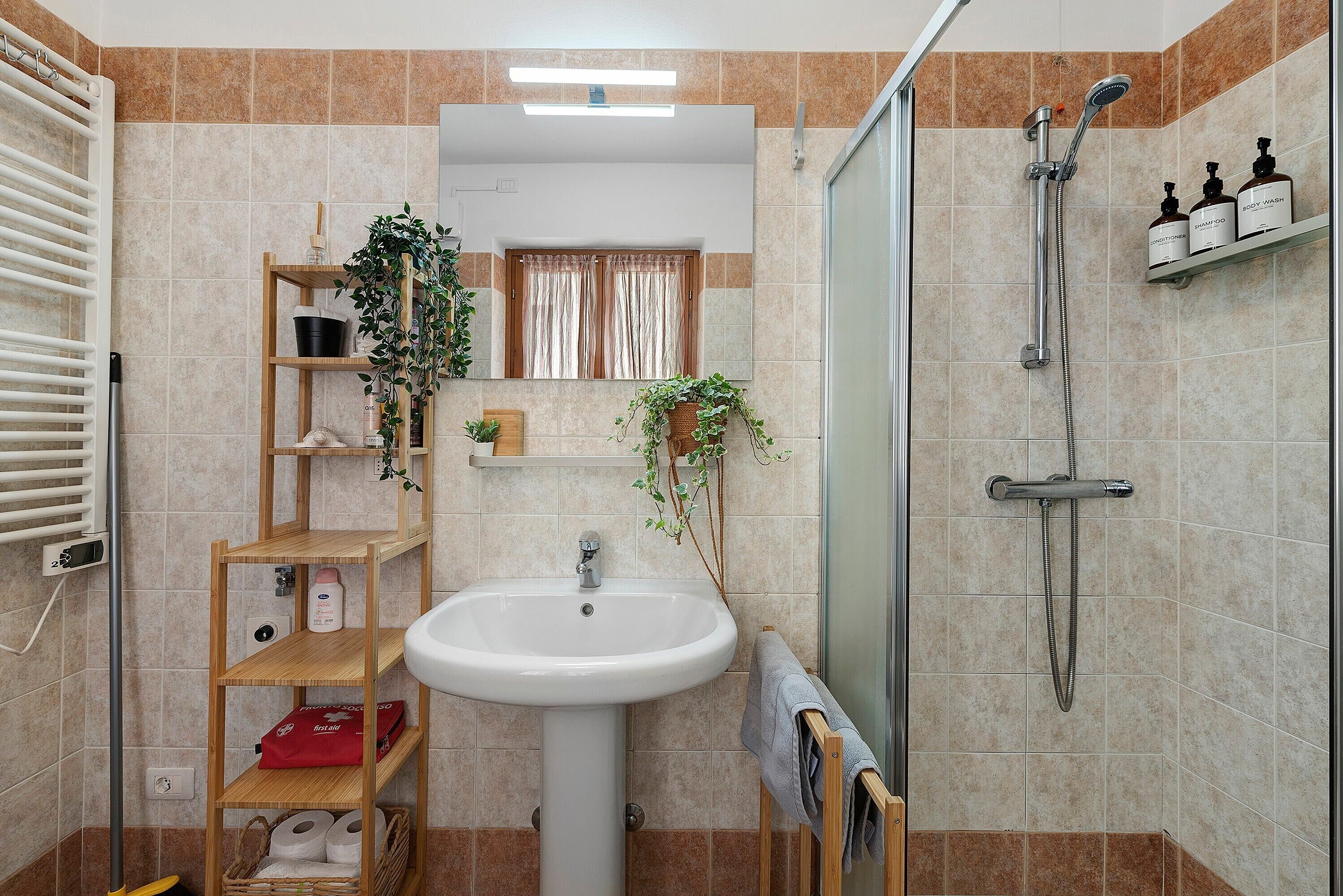 A bathroom with a sink, shower, shelves with toiletries, and a window with curtains.