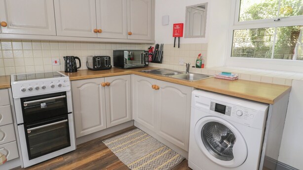 A kitchen with white cabinets, a stove, a washing machine, and a window with a view of trees. - Ysgoldy (Dolwyddelan)