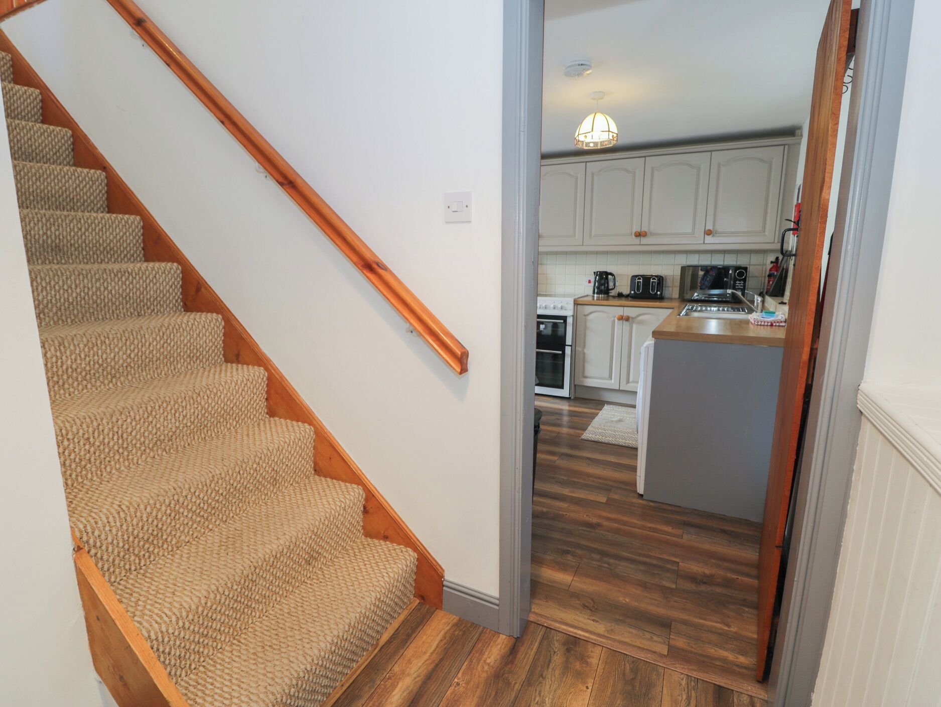 Una escalera conduce a una cocina con piso de madera, gabinetes blancos y una lámpara colgante.