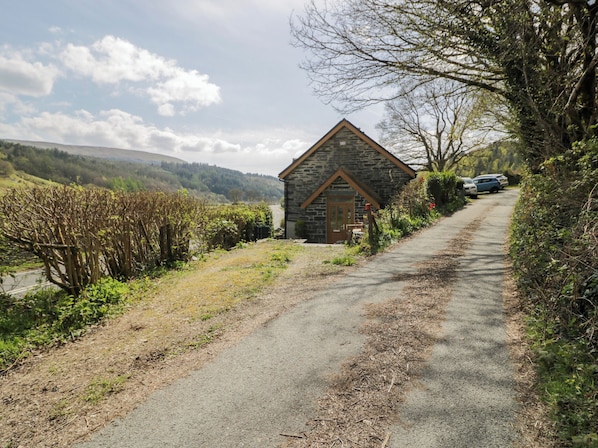 A stone building with a triangular roof, a paved path, and a vehicle parked nearby. - Ysgoldy (Dolwyddelan)