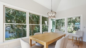 Un comedor con mesa y sillas de madera, ventanas amplias con vista a follaje, y una lámpara colgante.