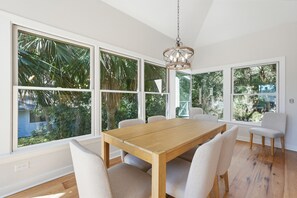 A dining area with a wooden table and chairs, large windows with a view of greenery, and a hanging light fixture. - 153 Mooring Buoy: Lowcountry Modern Home w/Private Pool, Walk to Beach (Hilton Head Island)