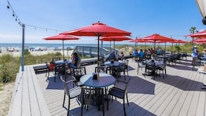 Una terraza al aire libre con mesas y sillas, sombrillas rojas y vista a la playa.