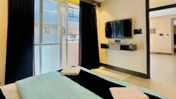 Ett hotellrum med en säng, en takfläkt, en tv som är monterad på väggen och ett fönster med gardiner.
