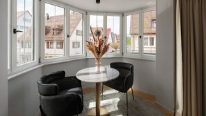 Habitación de esquina con ventanas amplias, una mesa redonda, dos sillas y un florero con plantas secas.