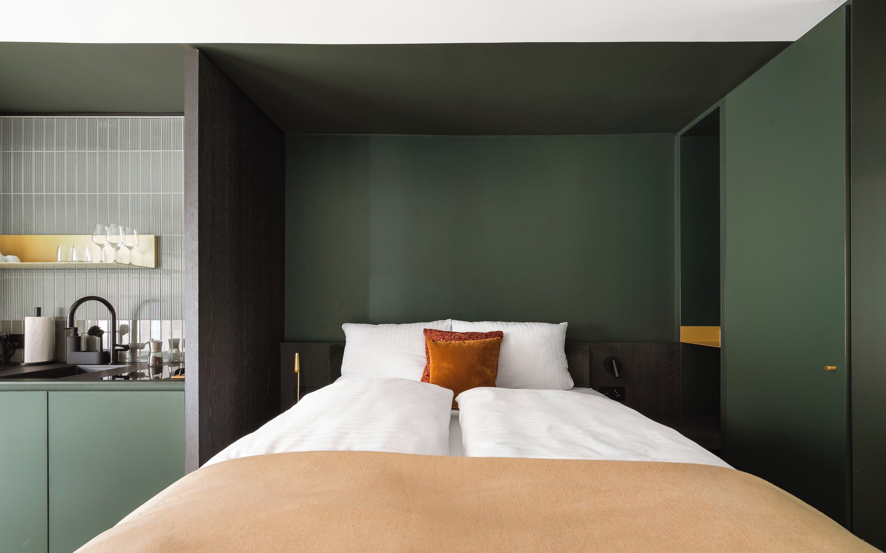 Une chambre d’hôtel moderne équipée d’un lit, d’une table de chevet, d’un miroir et d’un coin cuisine intégré avec un évier et une machine à café.