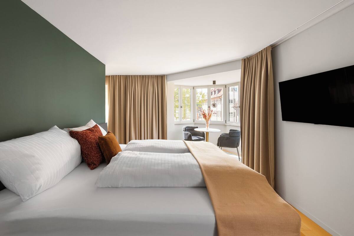 Ein Hotelzimmer mit Bett, Fernseher, Fenster mit Vorhängen und Blick auf Gebäude.