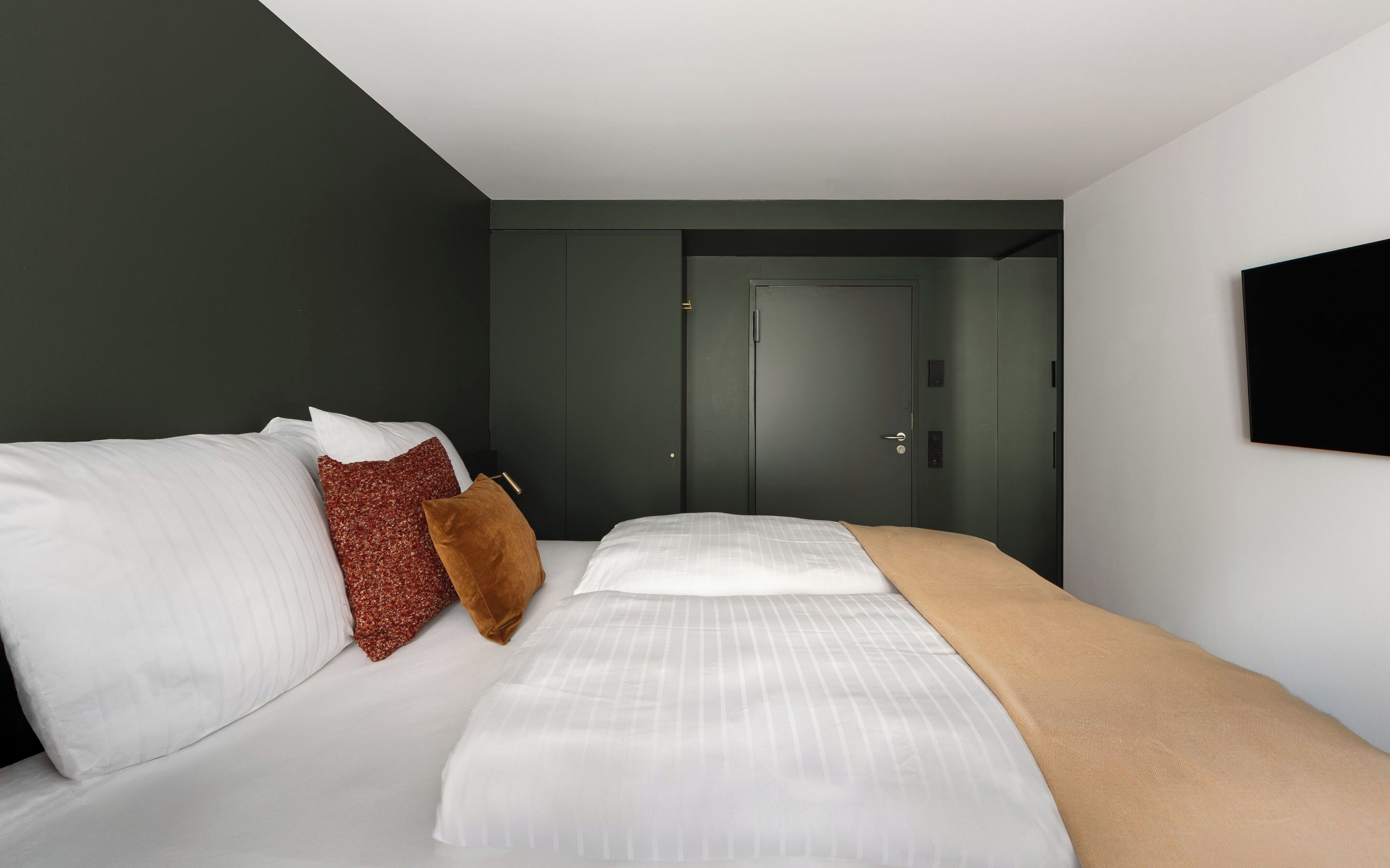 A modern hotel room with a bed, a door, and a TV mounted on the wall.