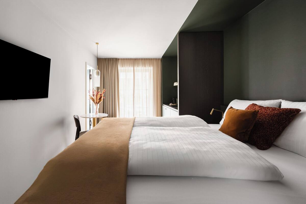 Ein modernes Hotelzimmer mit Bett, Schreibtisch, Stuhl, Fernseher und einem Fenster mit Vorhängen.
