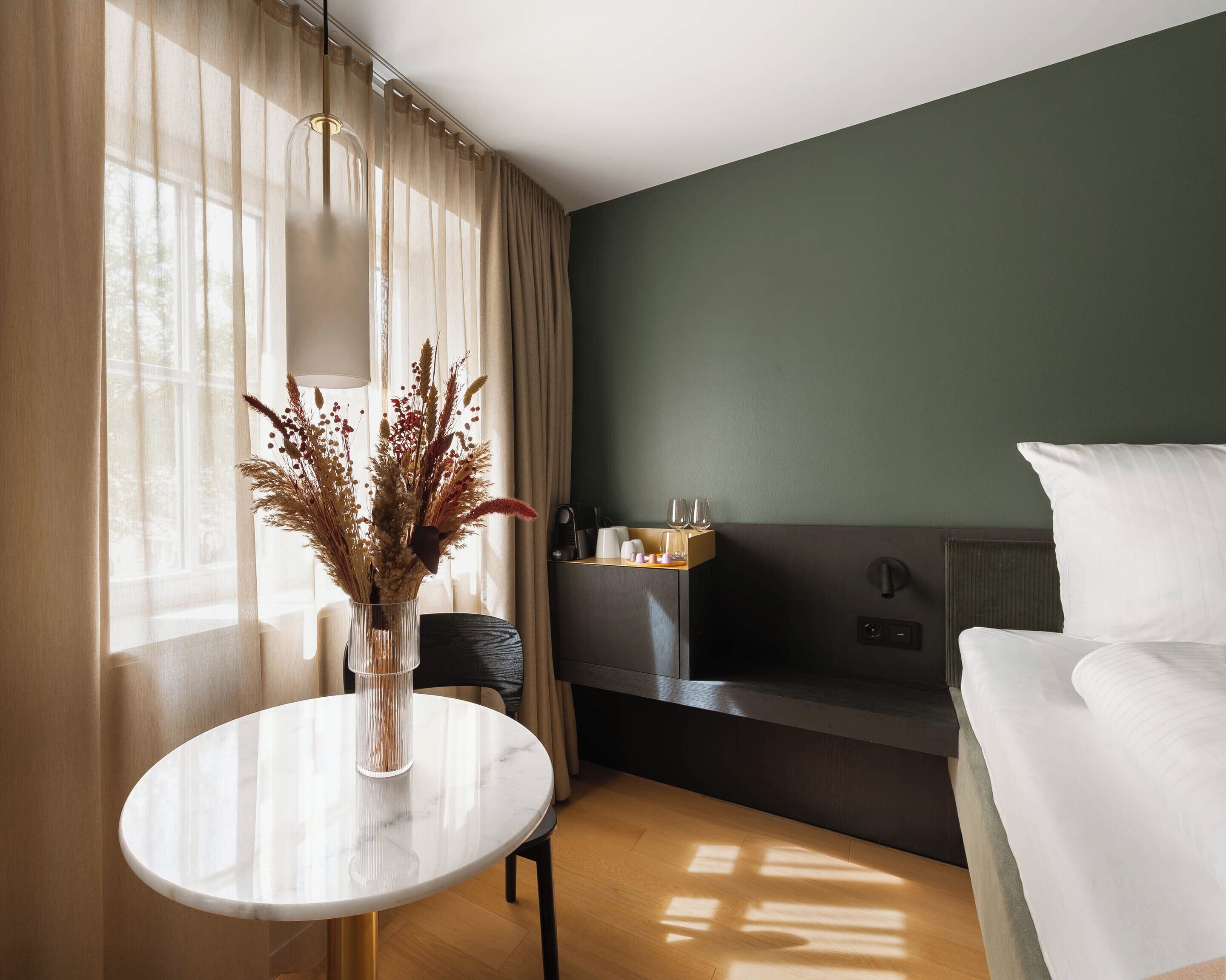 Habitación de hotel con cama, una mesita con un florero, un sofá, una cafetera y una ventana con cortinas.