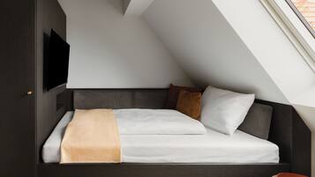 A modern loft-style bedroom with a sloped ceiling, a built-in TV, a bed with a headboard, and a small bedside table.
